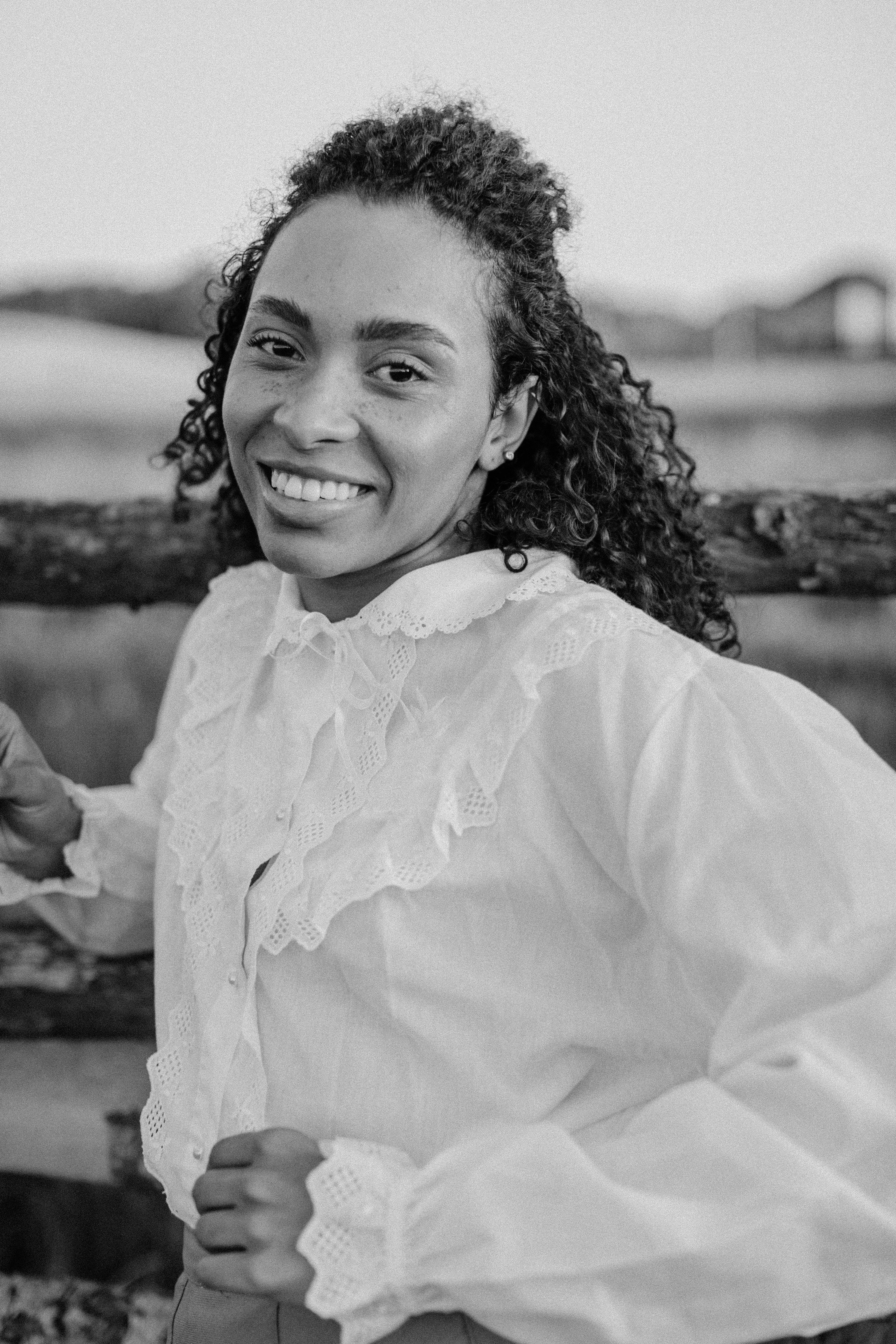 A smiling woman with curly hair in a vintage lace blouse, captured outdoors in black and white.