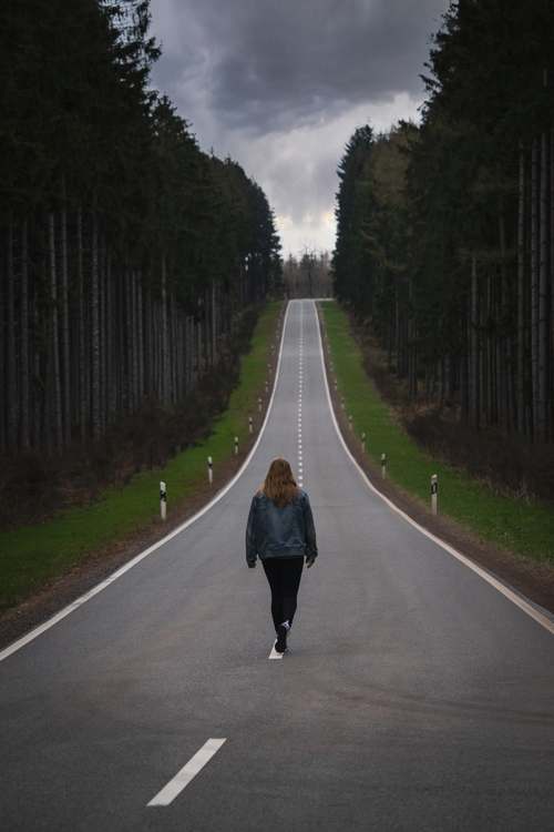Free A lone woman walks on a serene forest road beneath a moody, cloudy sky. Captured from behind. Stock Photo