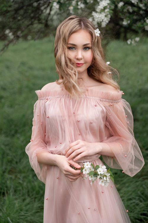 Free A young woman in a pink dress holding flowers, smiling in a lush outdoor setting. Stock Photo