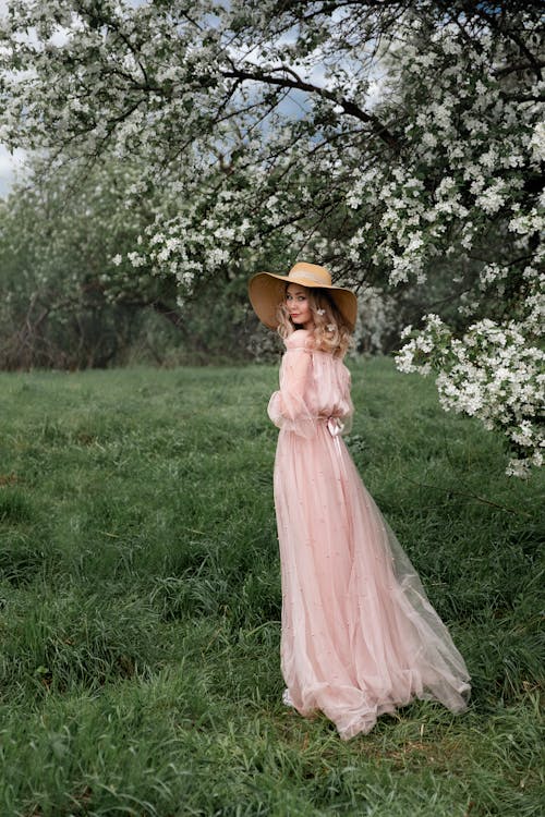A Woman in Pink Dress and Brown Hat Standing on a Green Grass Field
