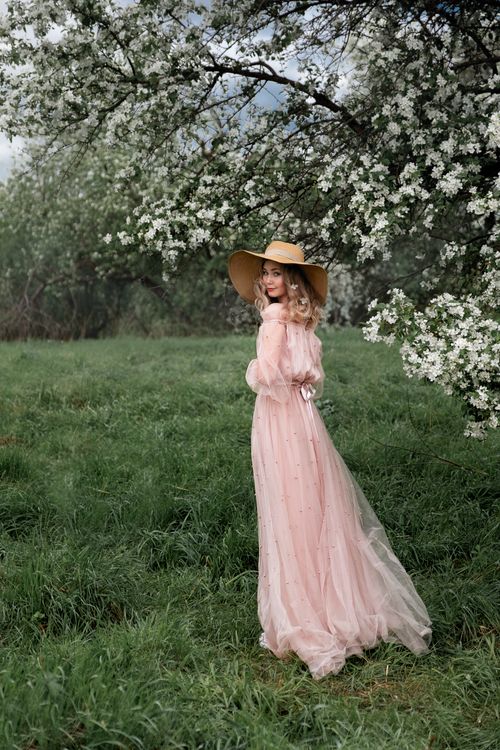 Free A woman in a pink dress and hat stands in a lush orchard with white blossoms, exuding grace. Stock Photo