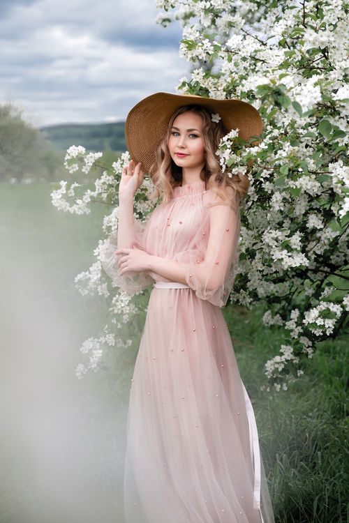 Free A smiling woman in a light pink dress and wide-brimmed hat stands amidst white blossoms in a springtime orchard. Stock Photo