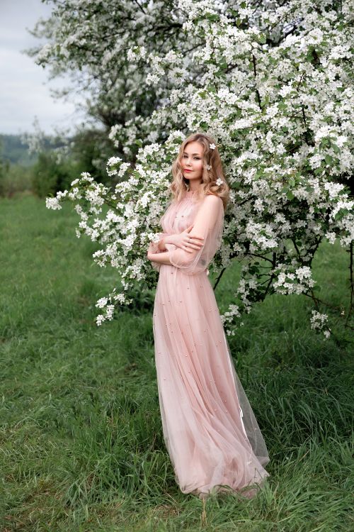 Free A woman in a flowing pink dress stands gracefully in front of a blooming tree in a lush green meadow. Stock Photo