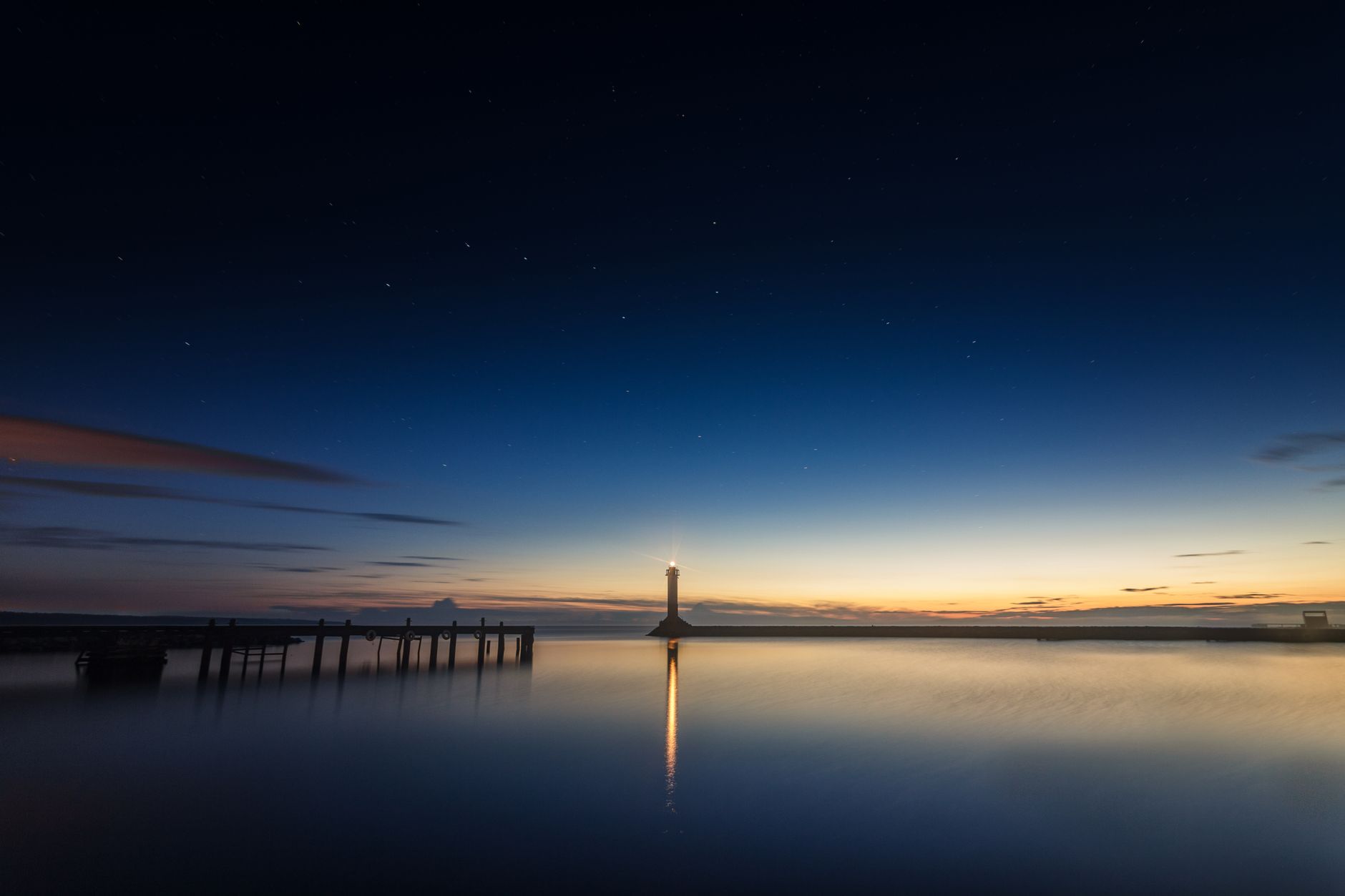 A tranquil twilight scene with a lighthouse reflecting on calm ocean waters, framed by a dock.