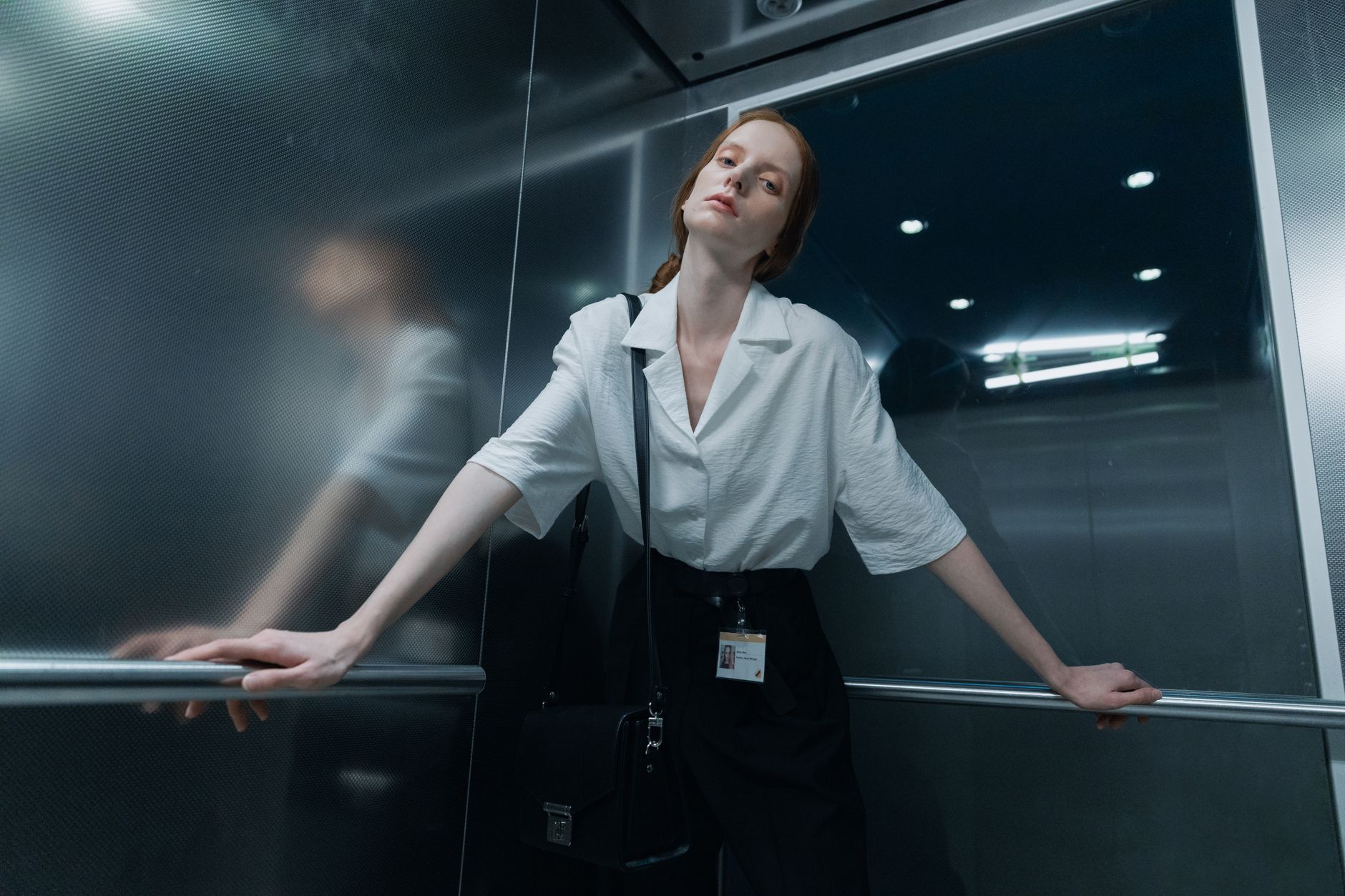 Confident woman standing in a modern elevator, wearing a white shirt and black pants, holding onto handrails.