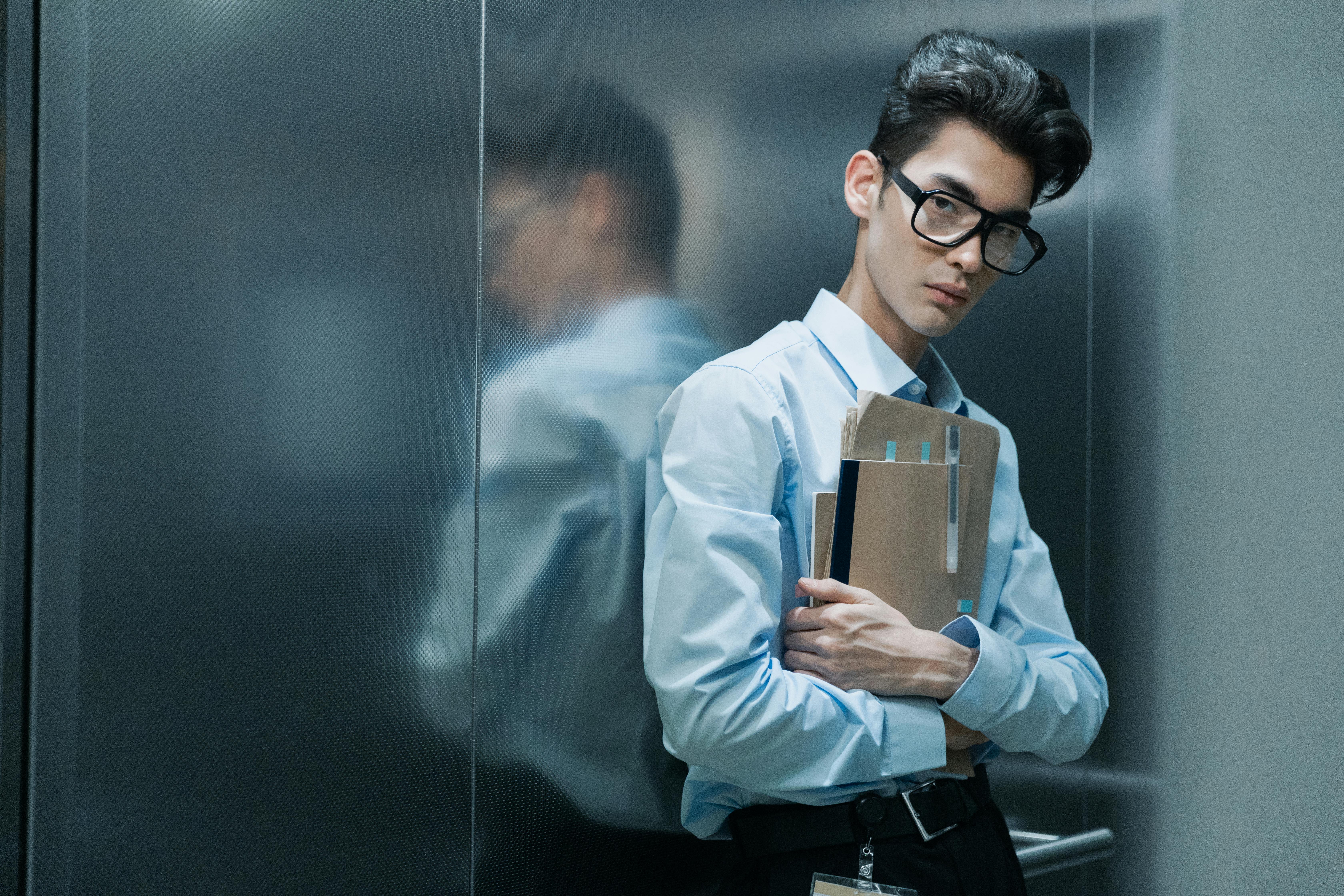 Asian man in glasses, blue shirt, holding papers inside an elevator.