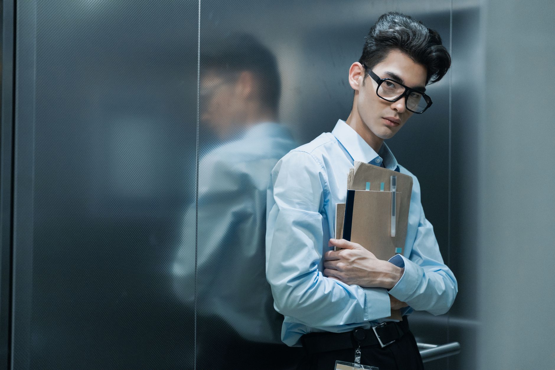 Asian man in glasses, blue shirt, holding papers inside an elevator.