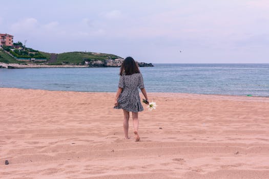 A woman in a short dress walks barefoot on the beach, holding lilies, under a cloudy sky.