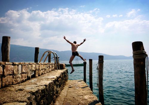 Free Man enjoying a carefree leap into a scenic Italian lake from a rustic stone pier, symbolizing freedom. Stock Photo