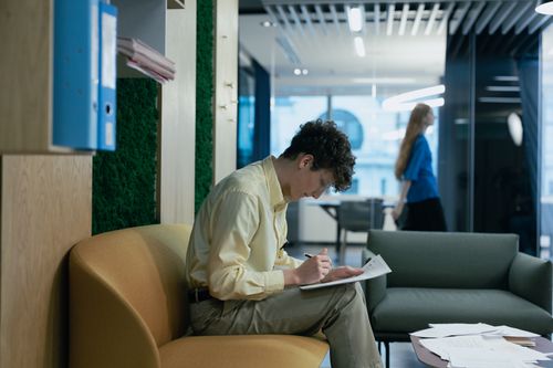 Free Office worker in yellow shirt writing notes on a couch, modern workspace with another person in the background. Stock Photo