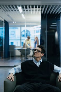 A thoughtful businessman sits in a modern office with glass panels, looking over his shoulder.