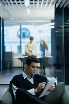 An adult man taking notes in a modern office setting with colleagues working in the background.