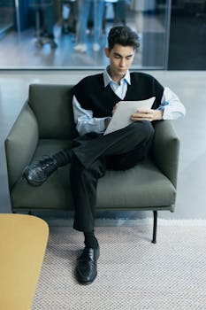 A young man in business attire, seated, reviewing documents in a modern office setting.