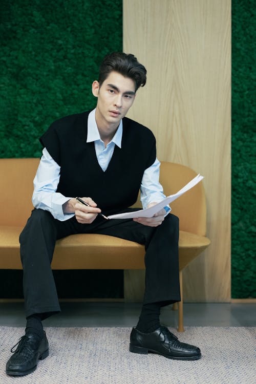 Man in Black Vest Sitting on Brown Wooden Bench Holding a PPaper