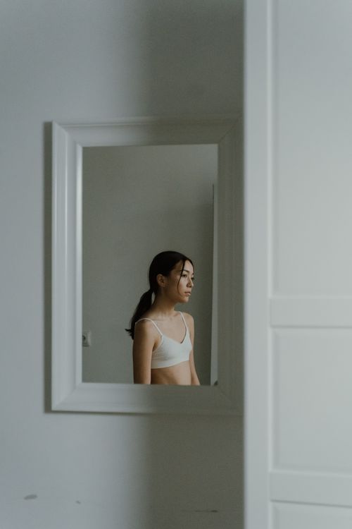 Free A woman in a mirror reflection stands softly lit in a minimalist white room. Stock Photo