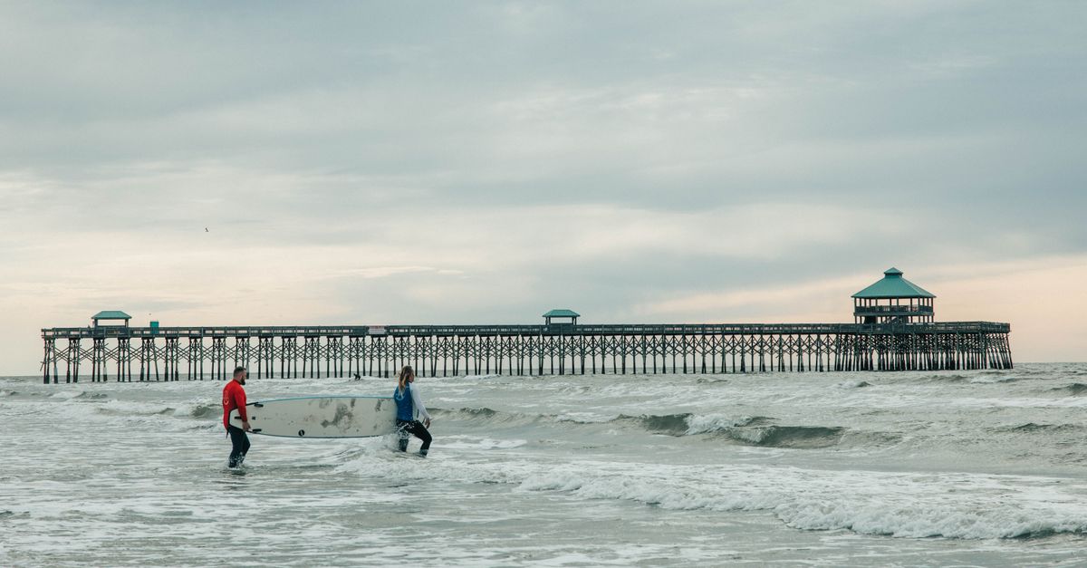 Two surfers carrying boards near Folly Beach Pier, SC, on an overcast day.