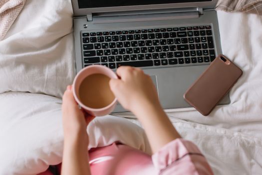 A cozy morning scene with a woman holding coffee, phone, and laptop on bed.