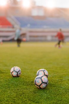 Three soccer balls on grass field with blurred players practicing in the background.
