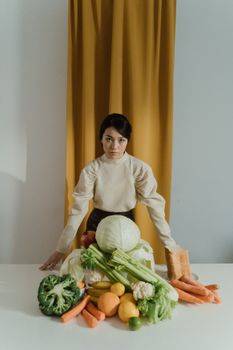 A woman stands behind a table of assorted fresh vegetables against a mustard curtain.
