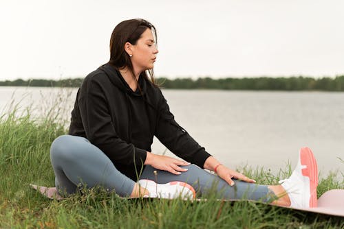 Free Woman in Black Shirt and Gray Pants Sitting on Green Grass Field Near Body of Water Stock Photo