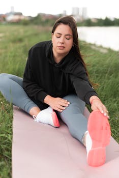 Woman in outdoor setting stretching on a yoga mat by a river, focused on wellness.
