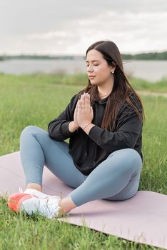 A brunette woman meditating on a yoga mat in a grassy outdoor setting for wellness.