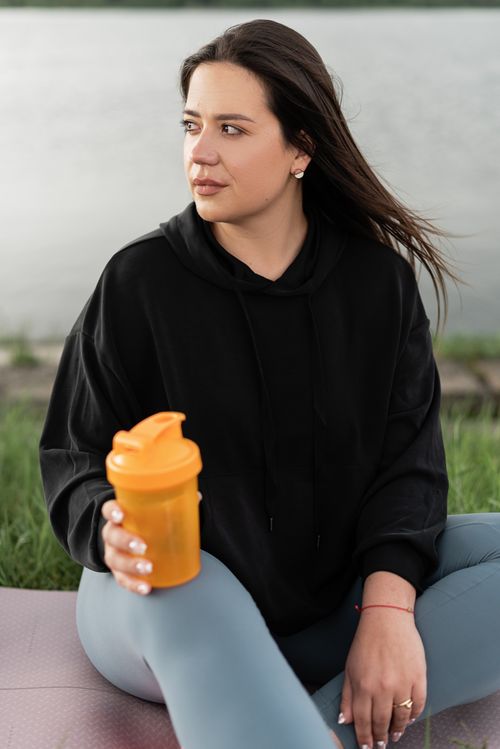 Free Woman in black hoodie holding an orange shaker bottle, sitting on a yoga mat by the water. Stock Photo