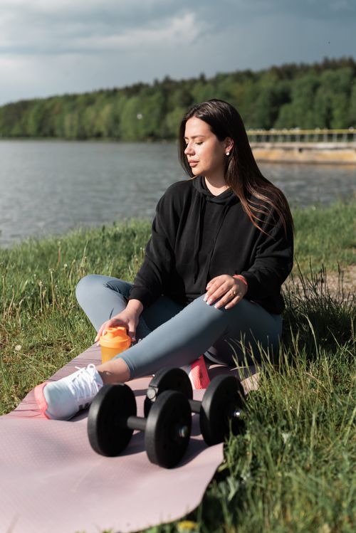 Free Woman sitting on yoga mat with dumbbells by a scenic lake, enjoying outdoor exercise. Stock Photo
