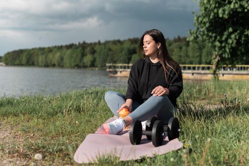 Free Woman sitting on yoga mat beside a lake with exercise equipment, enjoying a calm outdoor workout. Stock Photo