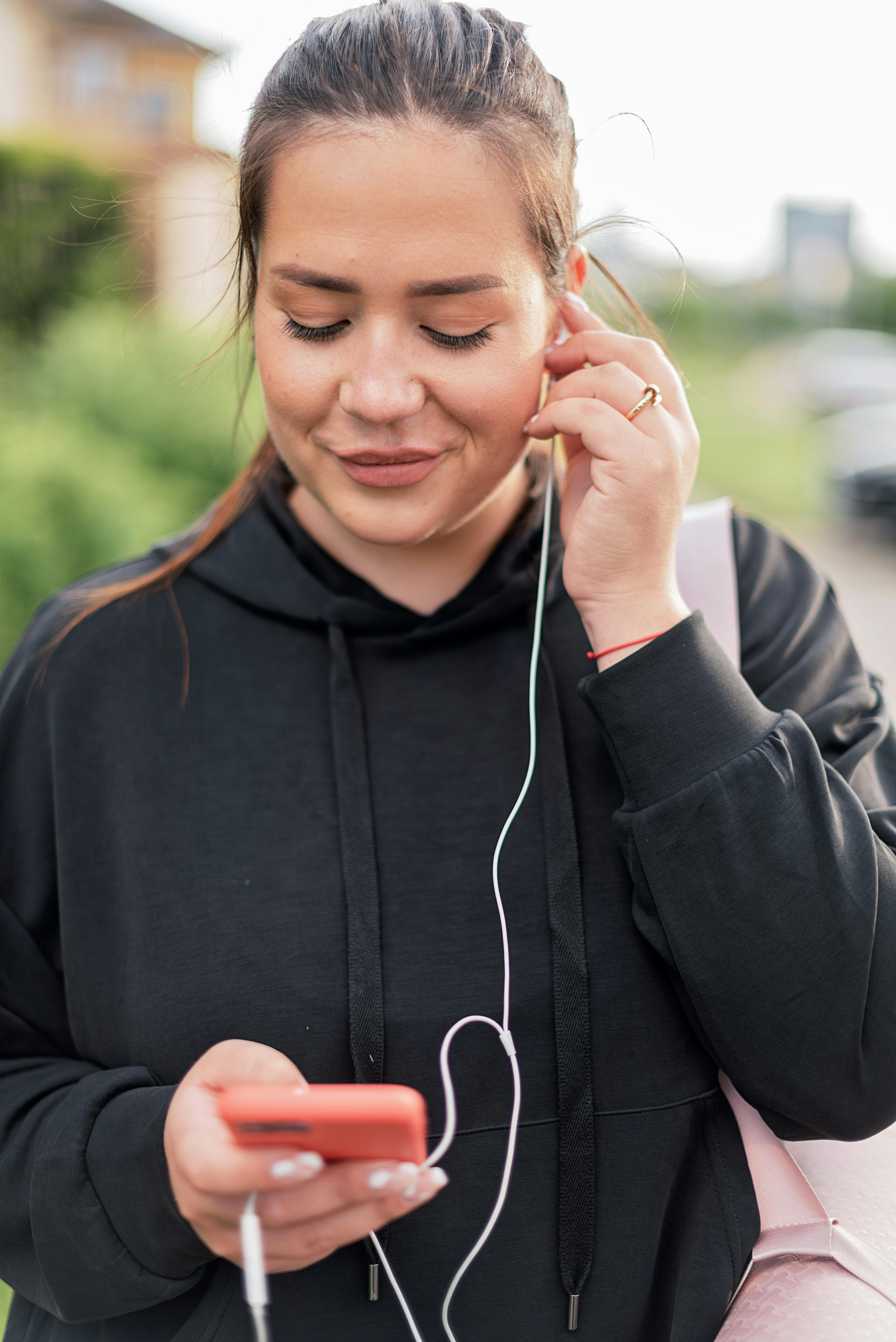 A woman in a black hoodie listens to music using headphones and a smartphone outdoors.