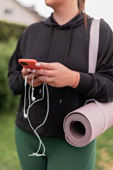 Woman holding a smartphone and yoga mat outdoors, ready for a workout.
