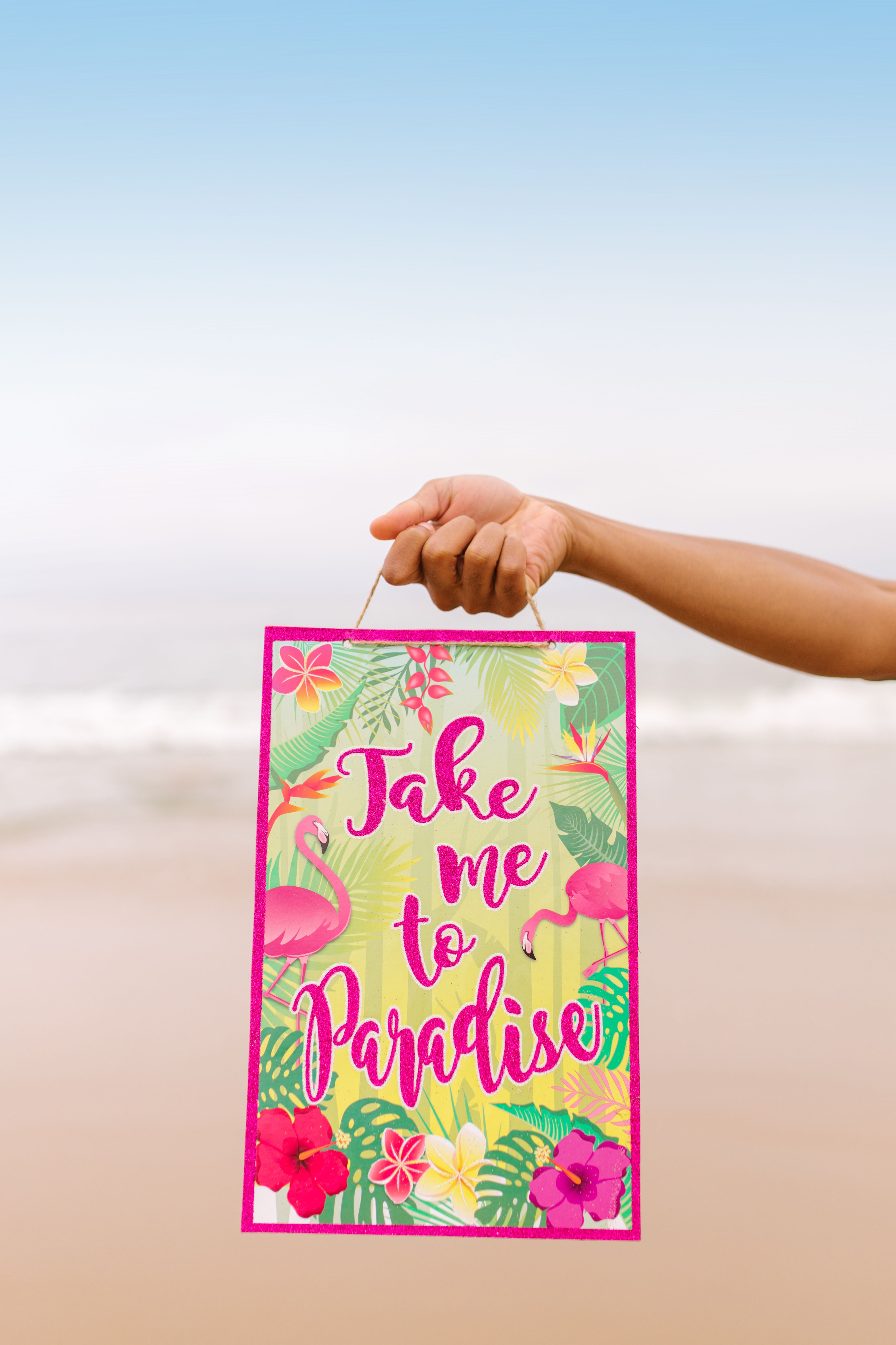 A hand holds a colorful tropical-themed poster on a sunny beach, inviting to paradise.