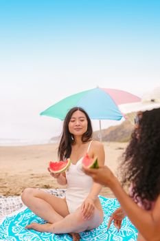 Two women relaxing on a beach under an umbrella, eating watermelon on a sunny summer day.