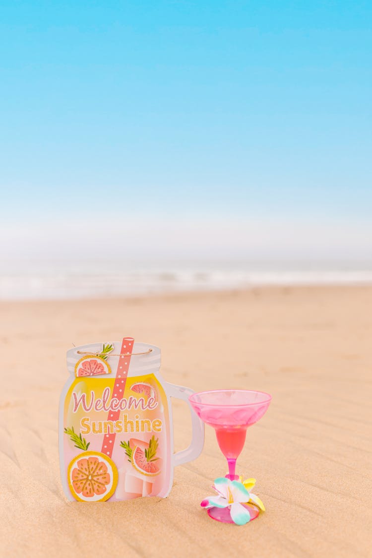 Pink Cocktail Glass And Colorful Postcard On Sand By Sea Shore