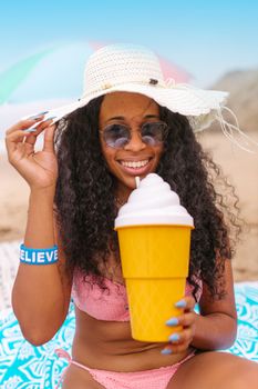 Happy woman wearing a sun hat and sunglasses, enjoying a beach day with a large ice cream themed drink.
