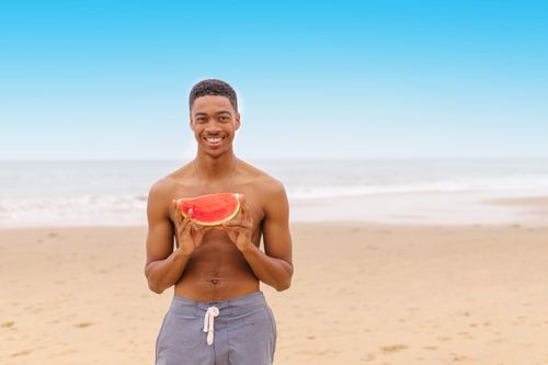 Free Shirtless man smiling and holding a watermelon on a sunny beach day. Stock Photo