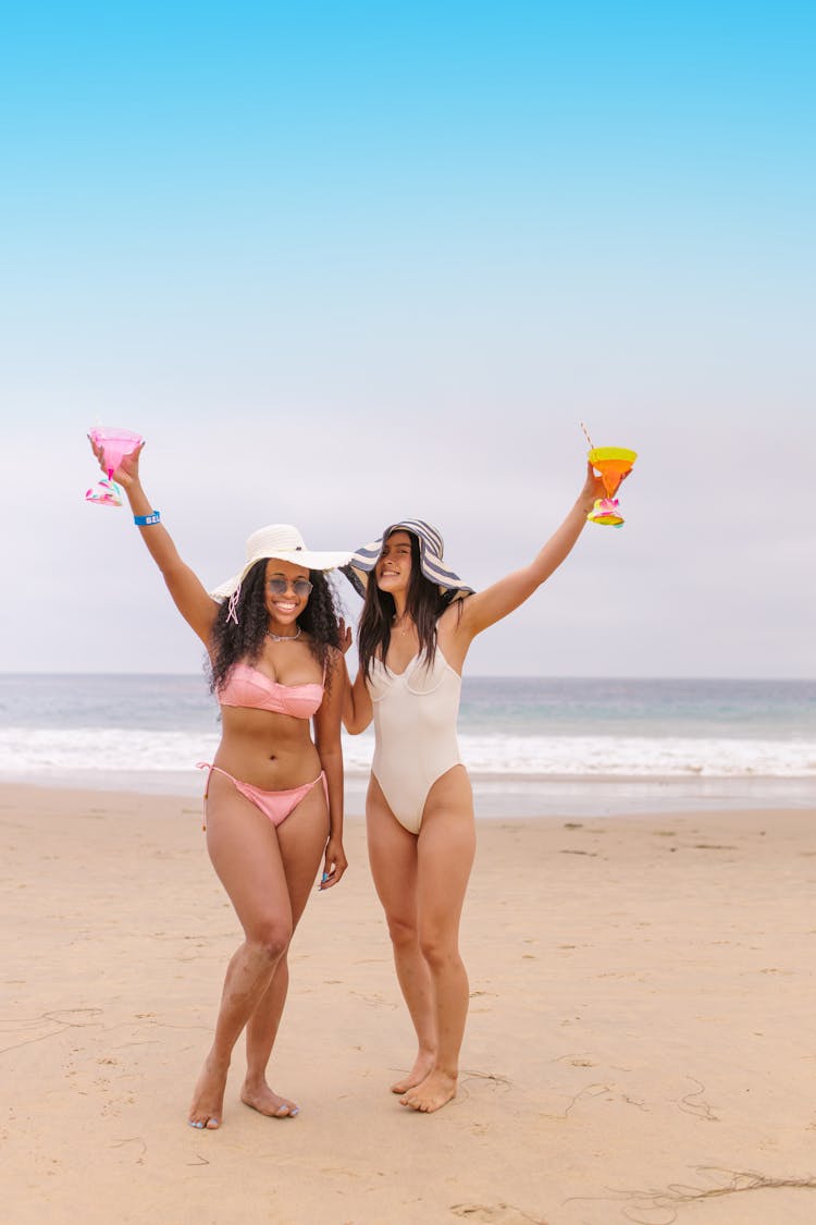 Women In Sexy Bikinis Standing On The Shore While Holding Glasses Of Juice