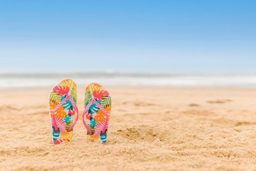 Vibrant patterned flip flops standing on a sandy beach with a clear blue sky.