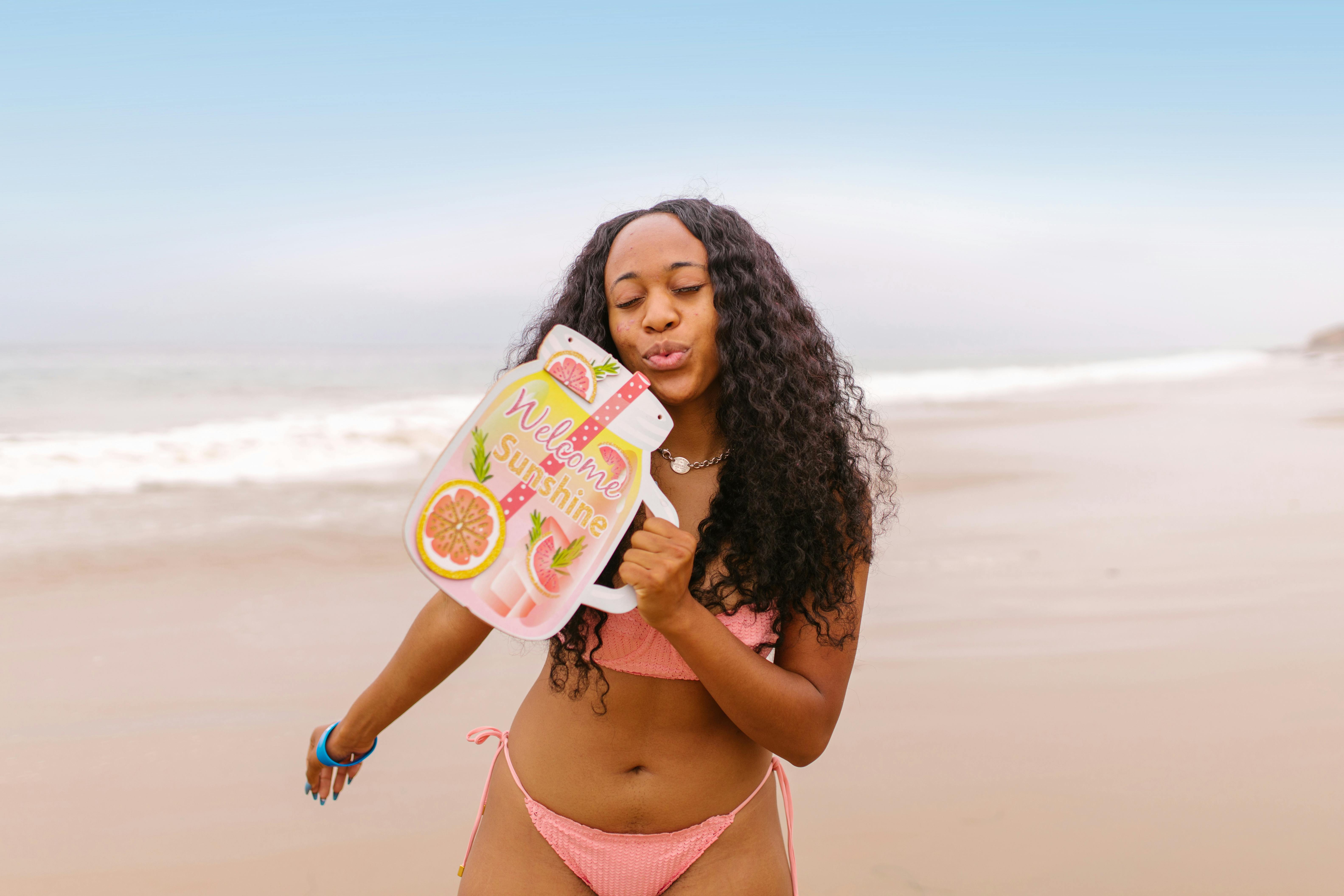 A woman in swimwear enjoys a sunny beach day, holding a refreshing drink sign that says 'Welcome Sunshine'.