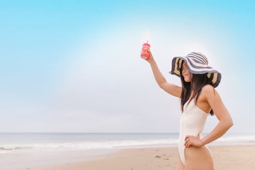 A Woman in White Swimsuit Standing on the Shore while Holding a Glass of Juice