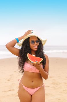 African American woman in a pink bikini enjoying watermelon on a sunny beach.