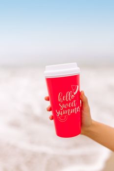 A vibrant red cup with 'hello sweet summer' message held over a beach background.