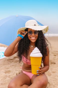 Woman in bikini and hat smiling on beach under umbrella holding a large cup.