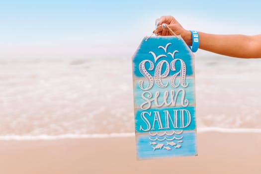 A beach sign reading 'Sea Sun Sand' held by a hand with a bracelet, capturing summer vibes.