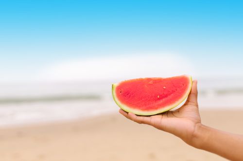 Free A hand holding a slice of watermelon against a beach background with blue sky. Stock Photo
