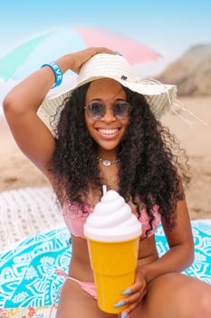 Joyful woman in a pink bikini and summer hat holding a large ice cream cup on the beach.