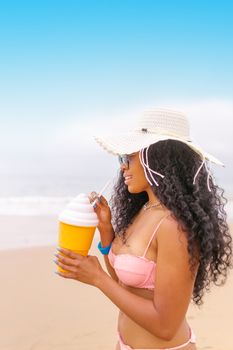 Smiling woman in a bikini with a sun hat enjoying a refreshing drink by the beach.