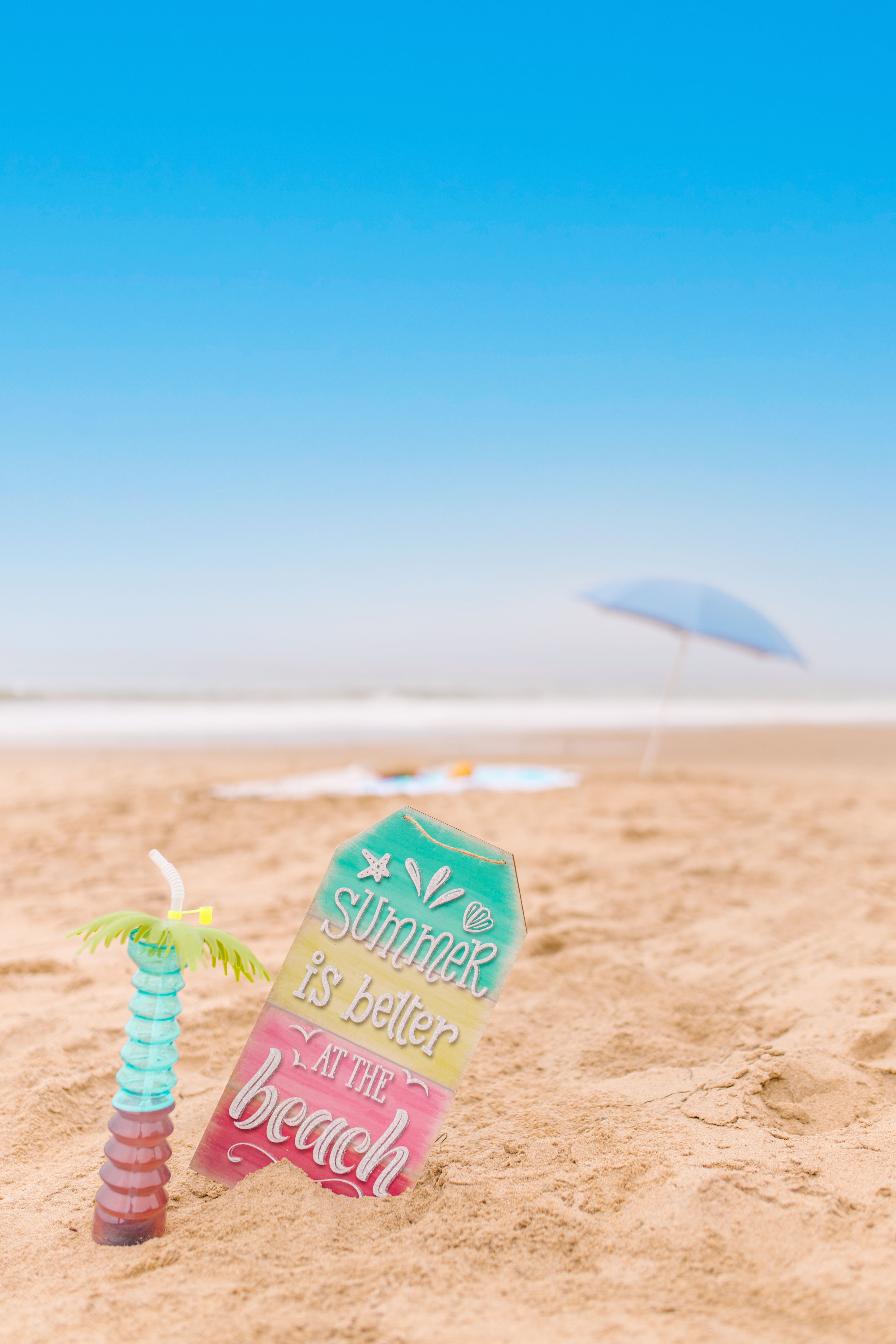 Vibrant beach sign and drink capture summer vibes on sandy beach with blue sky.