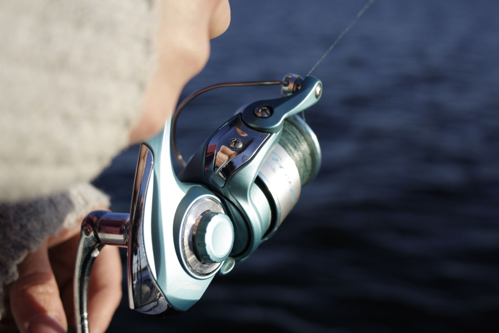 Detailed shot of a person's hand holding a fishing reel by the water, illustrating outdoor fishing activities.