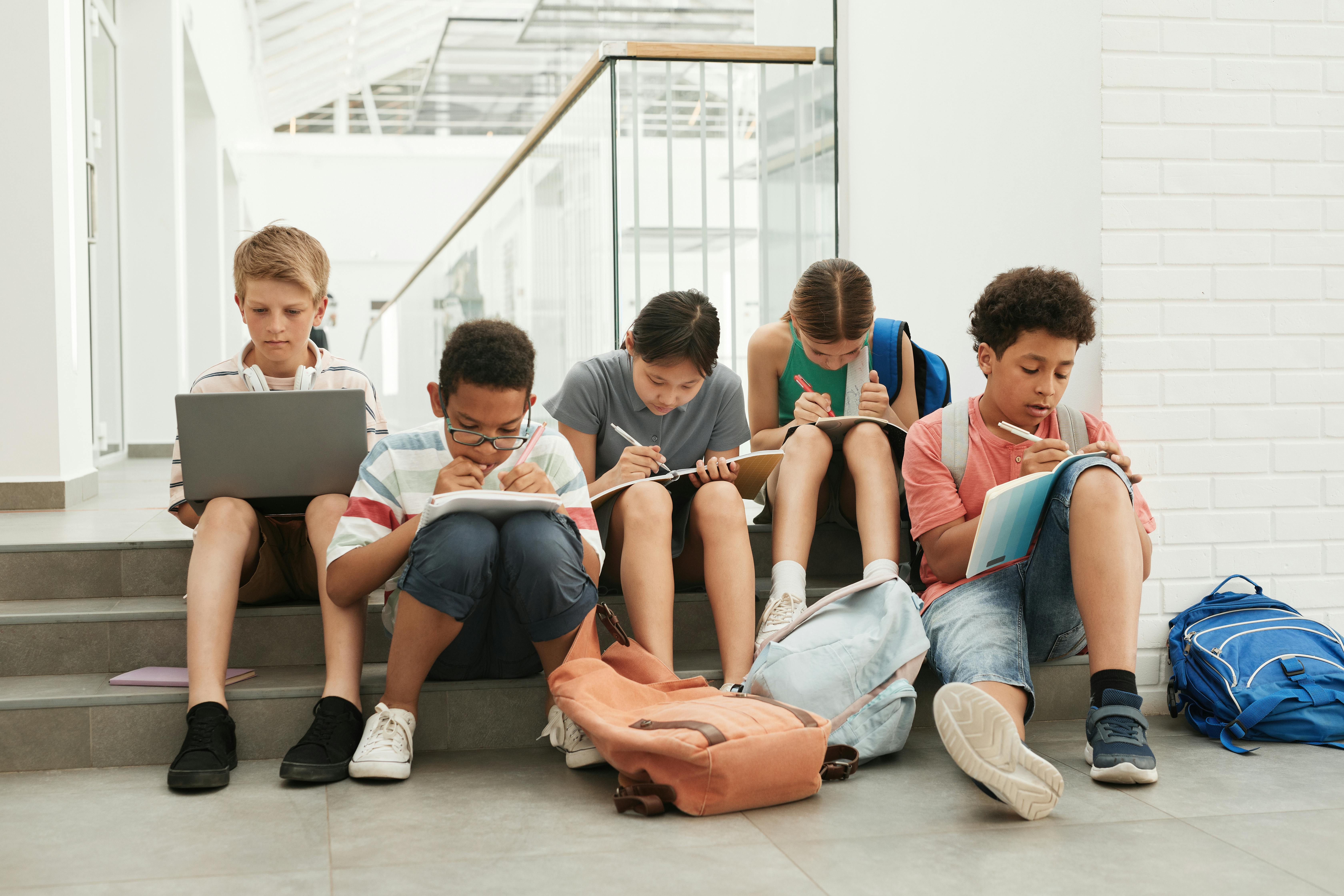 A group of school students sitting in a hallway, focused on studying and writing.